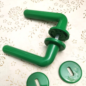 Peut inclure: Deux poignées de porte vertes avec des rosaces rondes assorties. Les poignées sont en forme de L avec une poignée arrondie et une base circulaire. Les rosaces ont une forme de trou de serrure. Les articles sont posés sur un fond à motifs.