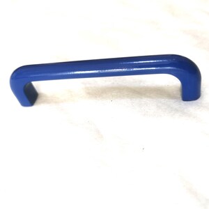 Op de afbeelding: Een helderblauwe handgreep met een gebogen ontwerp. De handgreep is gemaakt van een solide materiaal en staat tegen een witte achtergrond. De handgreep is een hardwarecomponent, waarschijnlijk voor een kast of lade.