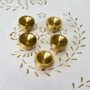 May include: Five gold-toned metal knobs with a rounded top and a small stem. The knobs are arranged in a semi-circle on a white surface with a floral pattern.