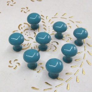 Puede incluir: Ocho pomos de armario de color azul turquesa con un acabado brillante. Los pomos están dispuestos en círculo sobre una superficie blanca con un patrón floral dorado.