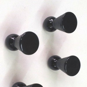 Op de afbeelding: Vier zwarte metalen haken met een ronde basis en een conische bovenkant. De haken zijn gemonteerd op een witte muur.