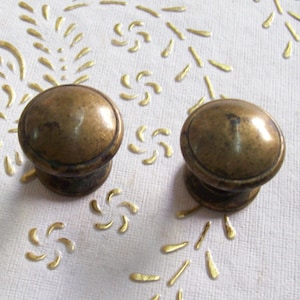 Puede incluir: Dos pomos de armario de latón antiguo con la parte superior redondeada y una base ligeramente elevada. Los pomos están sobre una superficie blanca con un patrón floral dorado.