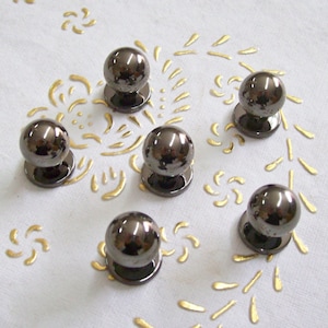 Puede incluir: Seis pomos de armario de metal negro con forma redonda ligeramente abovedada. Los pomos están dispuestos en semicírculo sobre una superficie blanca con un patrón dorado.