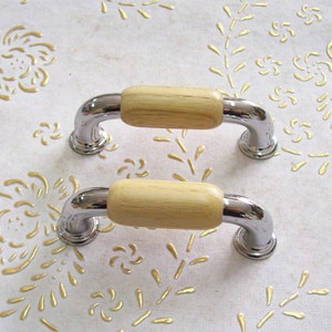 Könnte beinhalten: Zwei verchromte Schrankgriffe mit Holzgriffen. Die Griffe befinden sich auf einer weißen Oberfläche mit einem goldenen Blumenmuster.
