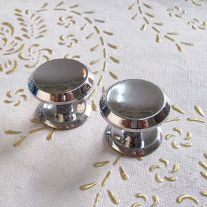 以下が含まれることがあります： 丸い、わずかに盛り上がったトップと、より小さい丸いベースを持つ、銀色のキャビネットノブが2つ。ノブは、金色の花柄の白い表面にあります。