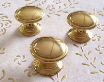 May include: Three gold-toned cabinet knobs with a round, slightly oval shape. The knobs have a raised, smooth surface and a small, round base.