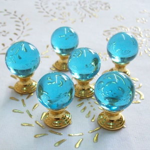 Può includere: Sei pomelli in vetro blu turchese con basi in metallo dorato. I pomelli sono rotondi e hanno una finitura liscia e lucida.