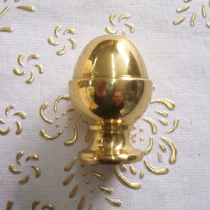 May include: A gold-colored egg-shaped decorative finial with a round base. The finial is made of metal and has a shiny finish.