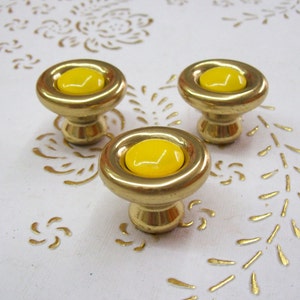 Könnte beinhalten: Drei goldene Schrankknöpfe mit gelben Mittelpunkten. Die Knöpfe befinden sich auf einer weißen Oberfläche mit einem goldenen Blumenmuster.