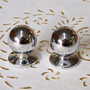 May include: Two silver-toned round cabinet knobs with a smooth, reflective surface. Each knob has a raised, circular base.