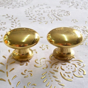 Könnte beinhalten: Zwei goldfarbene Schrankknöpfe mit runder Oberseite und zylindrischer Basis.