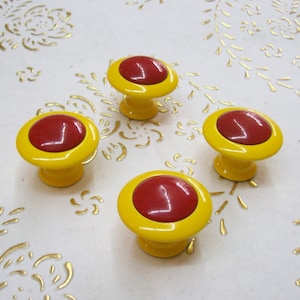 May include: Four yellow cabinet knobs with red centers. The knobs are arranged in a semi-circle on a white surface with a gold floral pattern.