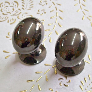 Op de afbeelding: Twee zwarte ronde kastknoppen met een glanzende afwerking. De knoppen bevinden zich op een wit oppervlak met een gouden patroon.