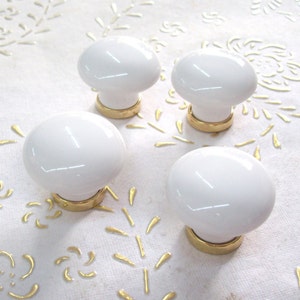 Peut inclure: Quatre boutons de cabinet en céramique blanche avec des bases en métal doré. Les boutons sont ronds et ont une finition lisse et brillante.