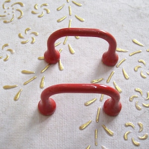 May include: Two red metal handles with a rounded top and a curved shape. The handles are on a white surface with a gold embossed pattern.