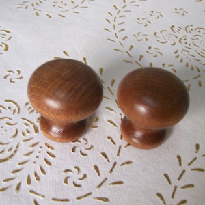 Può includere: Due pomelli in legno per mobili, con una parte superiore arrotondata e una base cilindrica corta. I pomelli sono di colore marrone caldo e sembrano essere in legno massello. Sono posizionati su uno sfondo bianco con un motivo floreale.