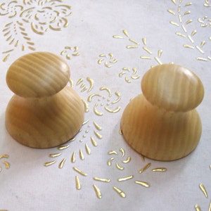 Op de afbeelding: Twee houten knoppen met een lichtbruine afwerking en een gegroefde textuur. De knoppen bevinden zich op een wit oppervlak met een gouden patroon.
