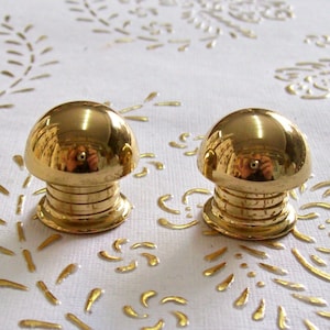 May include: Two gold-toned metal knobs with a rounded top and a ridged base. The knobs are on a white surface with a gold pattern.