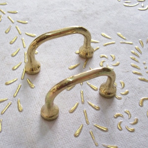 May include: Two gold-toned metal drawer pulls with a simple, curved design. The pulls are on a white surface with a gold pattern.