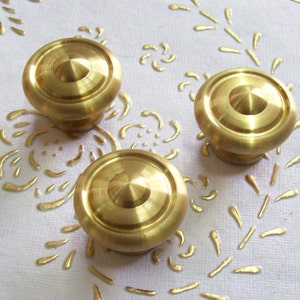 Op de afbeelding: Drie gouden metalen kastknoppen met een verhoogd middenstuk en een gladde, glanzende afwerking.