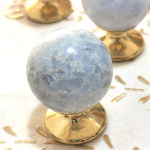 May include: Close-up of three blue crystal spheres on gold-colored pedestals. The spheres have a cloudy, textured appearance. The pedestals are shiny and reflective. Decorative gold leaf shapes are scattered around the bases.