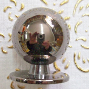 May include: A silver-toned round knob with a smooth, reflective surface. The knob is mounted on a circular base.