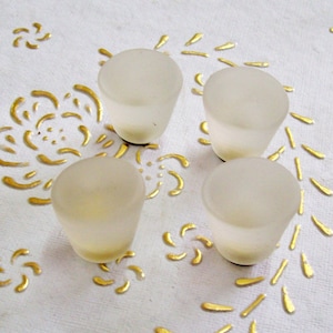 May include: Four frosted glass candle holders in a white, round shape. The candle holders are arranged in a square pattern on a white surface with a gold floral design.