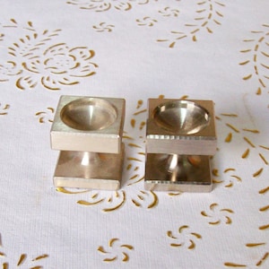 Peut inclure: Deux outils de tampon en métal argenté avec des bases carrées et un dessus rond et enfoncé. Les outils sont sur une surface blanche avec un motif floral doré.