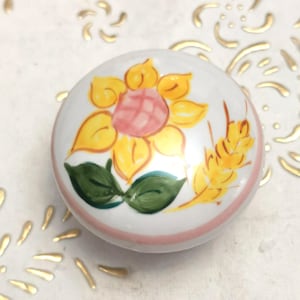Può includere: Pomello in ceramica bianca con un motivo a girasole dipinto a mano. Il girasole ha petali gialli, un centro rosa e foglie verdi. Una fascia rosa circonda la base del pomello e una spiga di grano è anch'essa dipinta.