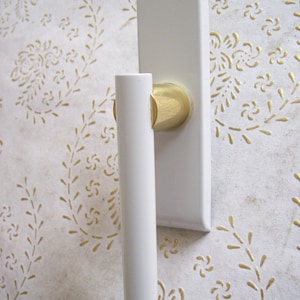 Op de afbeelding: Witte raamgreep met een gouden accent. De greep is bevestigd aan een wit rechthoekig plaat.