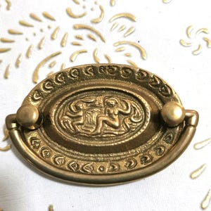 Op de afbeelding: Een ovale kastknop in goudkleur met een decoratief ontwerp met twee figuren in reliëf. De knop heeft een gladde, gepolijste afwerking en een eenvoudig, elegant ontwerp.