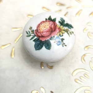 Pode incluir: Puxador redondo de cerâmica branca com um design floral. Apresenta uma grande rosa rosa, flores amarelas e azuis e folhas verdes. Este puxador decorativo é provavelmente para um móvel e tem um estilo vintage.