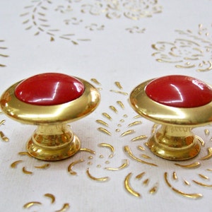 May include: Two gold cabinet knobs with red ceramic tops. The knobs are oval shaped with a raised rim around the red top.