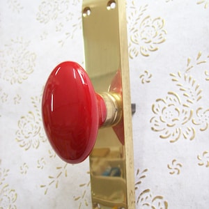 Może przedstawiać: Klamka drzwiowa wykonana z mosiądzu z jasnoczerwonym, owalnym gałką. Klamka ma dwa otwory montażowe.