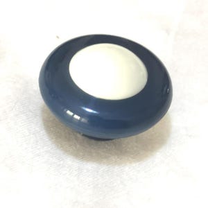Könnte beinhalten: Ein marineblauer und weißer Knopf mit glänzendem Finish. Der Knopf hat ein kreisförmiges Design mit einem weißen Zentrum und einem dunkelblauen Außenring. Das Objekt ist wahrscheinlich ein dekoratives oder funktionelles Hardwareteil.