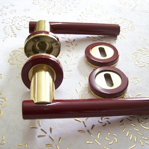 May include: A set of two door handles with a brass finish and burgundy red accents. The handles are cylindrical with a rounded end and come with two matching escutcheons.