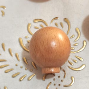 May include: A close-up of a light brown wooden knob. The knob is round and has a smooth surface. It is likely used for furniture or cabinets.