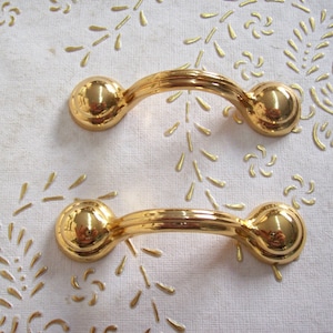 Könnte beinhalten: Zwei goldfarbene Schranktürgriffe mit abgerundeten Kanten und einem geschwungenen Design.