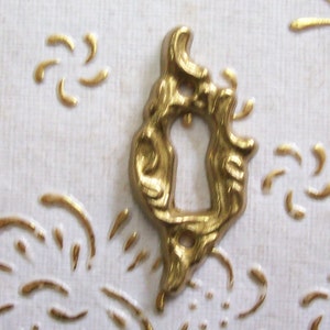 May include: A gold-coloured metal keyhole cover with an ornate design. The cover is shaped like a stylised letter D.