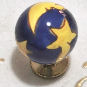 Op de afbeelding: Een decoratieve kastknop met een hemels ontwerp. De knop heeft een blauwe achtergrond met een gele maan en sterrenmotief. Hij is gemonteerd op een goudkleurige basis.