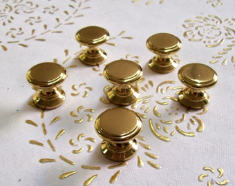 May include: Six gold-toned cabinet knobs with a round top and a cylindrical base. The knobs are arranged in a semi-circle on a white surface.