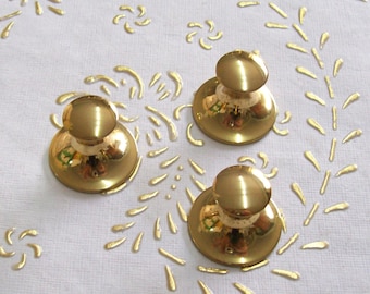 Puede incluir: Tres pomos de metal de color dorado con una parte superior redonda y una base plana. Los pomos están dispuestos en forma triangular sobre una superficie blanca.