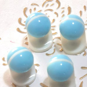 May include: Four blue and white striped glass thimbles. Each thimble features a light blue sphere with a white stripe, set on a white base. The thimbles are arranged on a white surface with gold decorative accents.