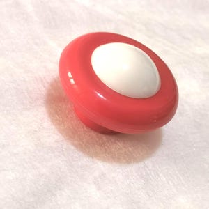 Peut inclure: Un bouton de meuble rouge et blanc de forme arrondie. Le bouton a un anneau extérieur rouge circulaire et un centre blanc légèrement bombé. Le bouton est probablement en plastique ou dans un matériau similaire.