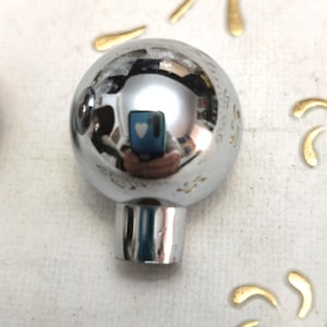 Könnte beinhalten: Eine glänzende, verchromte Kugel mit kurzem, zylindrischem Sockel. Die Kugel reflektiert die Umgebung. Goldfarbene, blütenblattförmige Akzente sind auf einer weißen Oberfläche um die Kugel herum verstreut. Es handelt sich wahrscheinlich um einen dekorativen Knopf oder Griff.