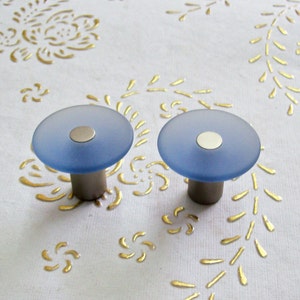 May include: Two frosted glass cabinet knobs with a silver base. The knobs are light blue and have a circular shape.