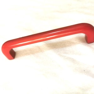 May include: A bright red, curved handle with a rectangular base. The handle is made of a solid material and has a smooth, glossy finish. The handle is designed for pulling or lifting and is likely used on a cabinet or drawer.