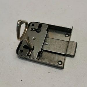 May include: A silver metal door lock with a key inserted into the keyhole. The lock has a rectangular shape and a silver finish.