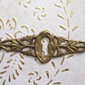 Könnte beinhalten: Ein goldfarbener Schlüsselschlitzabdeckung aus Metall mit einem verzierten Design. Die Abdeckung hat ein Schlüsselloch in der Mitte und ein dekoratives Muster auf beiden Seiten.