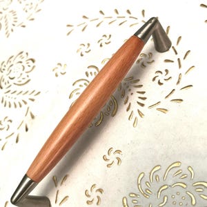 May include: A cabinet pull with a light brown wooden center and brushed nickel ends. The handle is set against a white surface with a gold floral pattern. The hardware is designed for easy installation.